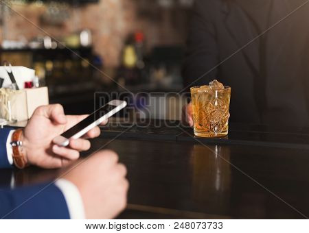 Bartender Giving Glass Of Whiskey To Customer In Bar, Businessman Using Mobile Phone, Copy Space