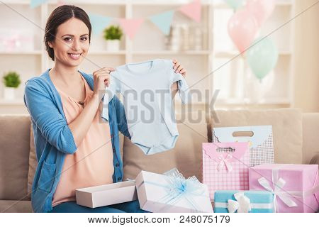 Smiling Pregnant Woman With Blue Baby Romper. Happy Pregnant Mother Holding Romper In Bedroom At Hom