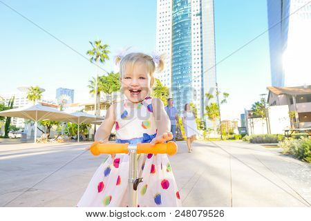 Emotional Cute Little Blondy Toddler Girl In Dress Riding Scooter In The City Park Recreation Area W