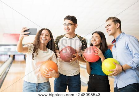 Smiling Teenage Girls And Boys Capturing Memories Of Bowling On Mobile Phone In Alley At Club