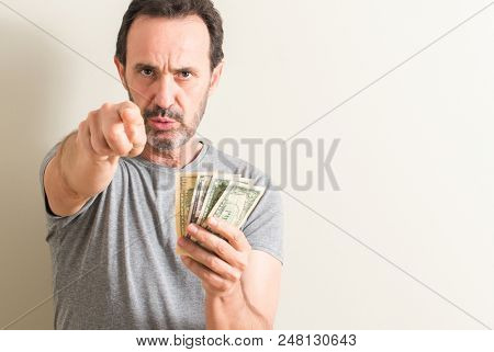 Senior man holding dollar money pointing with finger to the camera and to you, hand sign, positive and confident gesture from the front