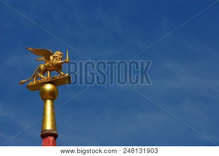 Saint Mark Winged Lion Golden Statuette Wielding A Sword Against Blue Sky, A Symbol Of The Old Venic