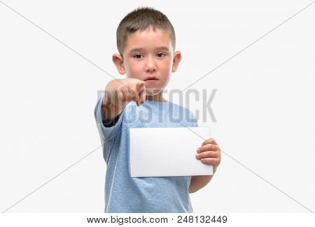 Dark haired little child holding a blank card pointing with finger to the camera and to you, hand sign, positive and confident gesture from the front