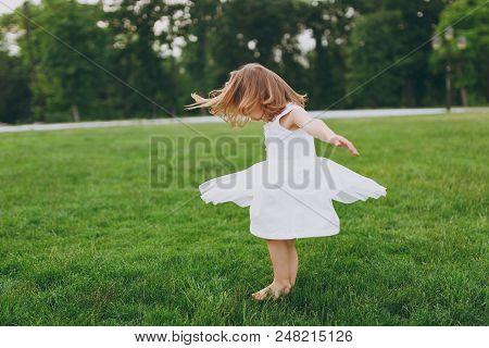 Pretty Little Cute Child Baby Girl In Light Dress Circling Around And Have Fun On Green Grass Lawn I