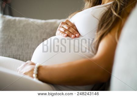 Close-up Of Pregnant Woman Relaxing And Sitting On The Side On The Sofa. Holding A Hands On The Tumm
