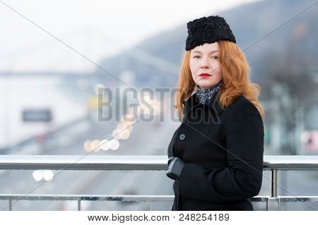 Aged Woman In Black Coat On Bridge. Urban Girl In Black Coat With Rouge Hair. Portrait Of Urban Woma