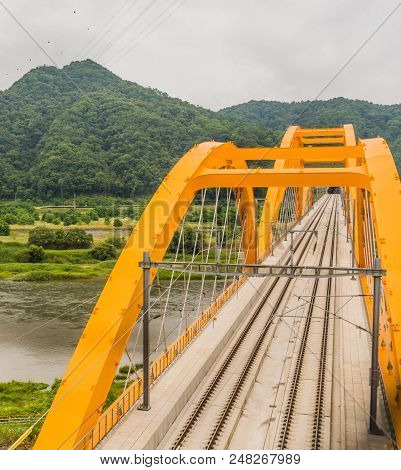 Top View Of Yellow Arched Truss Railroad Bridge Spanning A River In The Countryside On Cloudy Day.