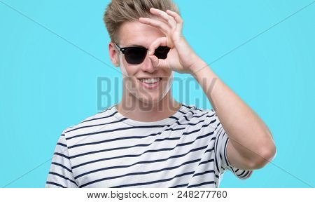 Young handsome blond man wearing sunglasess with happy face smiling doing ok sign with hand on eye looking through fingers