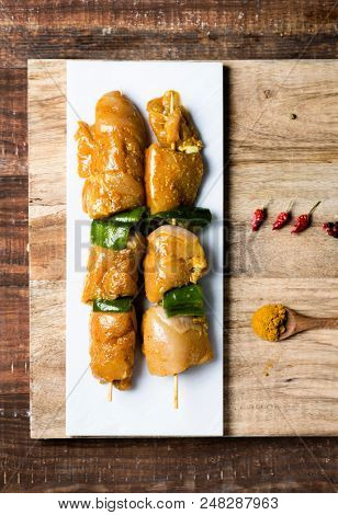 closeup of some raw spiced chicken meat skewers, mixed with green pepper, on a white marble tray, on a wooden table