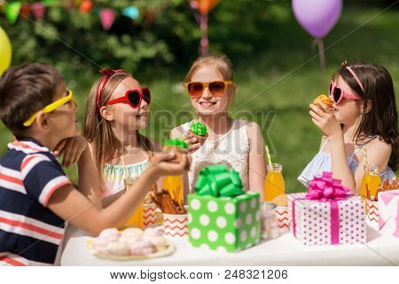 holidays, childhood and celebration concept - happy kids in sunglasses sitting at table on birthday party at summer garden and eating cupcakes