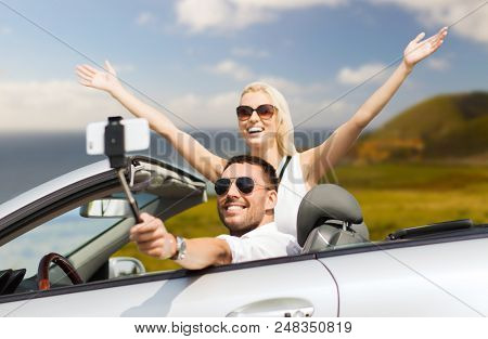 road trip, technology and travel concept - happy couple driving in convertible car and taking picture by smartphone on selfie stick