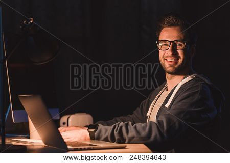 Handsome Young Programmer In Eyeglasses Smiling At Camera While Working With Laptop And Desktop Comp