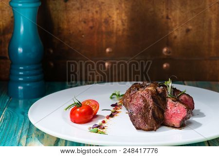 Close-up Appetizing Beef Steak With Herbs And Tomato On A White Plate, On A Wooden Table. Brown Back