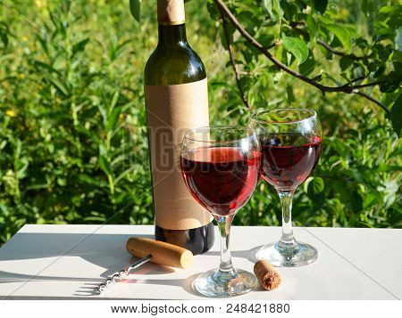 Red Wine Poured Into Wine Glass On Background Of Green Tree Leaf Foliage. Purpie Flower