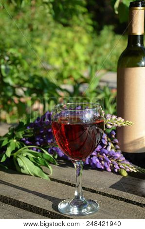 Red Wine Poured Into Wine Glass On Background Of Green Tree Leaf Foliage. Purpie Flower