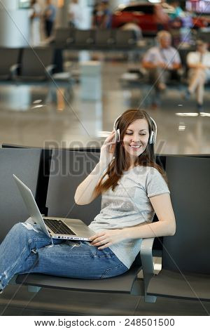 Young Smiling Traveler Tourist Woman With Headphones Listening Music Working On Laptop, Wait In Lobb