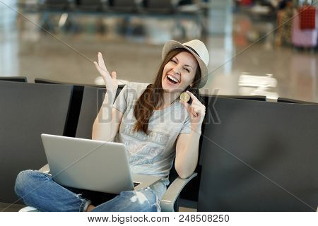 Young Laughing Traveler Tourist Woman In Hat Sitting, Working On Laptop, Holding Bitcoin, Spreading 