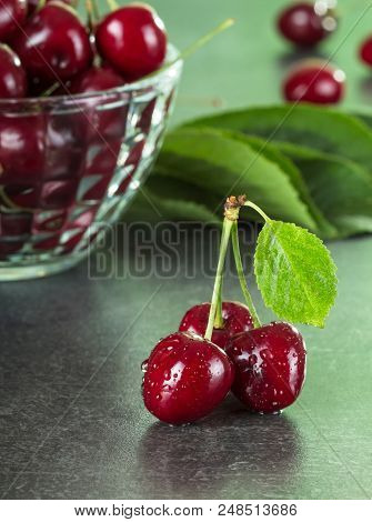 Three Sweet Berries In Drops Of Water, Next To Bowl Of Cherries, On Gray Surface