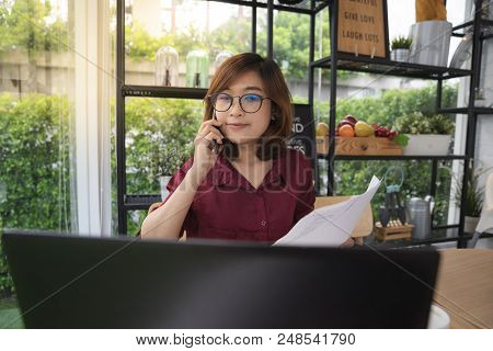 A Young Lady Wears Glasses Working On Laptop And Talking On A Mobile Phone In Cafe