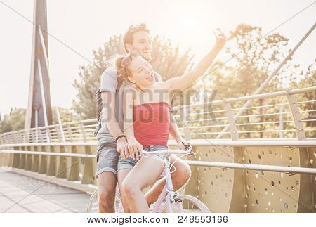Happy Millennial Couple Taking Photo Selfie With Smartphone On Bicycle - Young Students Having Fun T