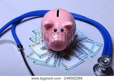Piggy Bank With Dollar Notes And Stethoscope On A Wooden Desk.
