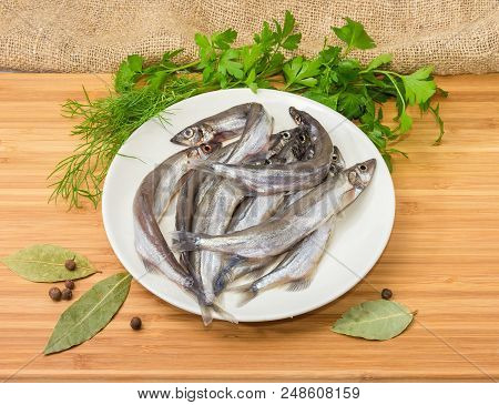 Defrosted Uncooked Capelins On The White Dish, Bay Leaf, Allspice And Fresh Parsley And Dill On A Wo