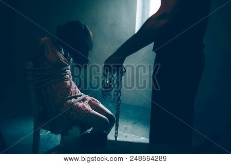 Dark Picture Of Kidnapper Standing Besides His Victim. He Is Holding Chains In Hand. Girl Is Sitting