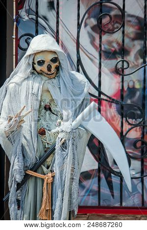 A Papier Mache Doll Depicting Death Stands Near A Wrought-iron Fence