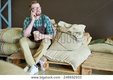 Full Length Portrait Of Handsome Young Man Sitting On Heap Of  Burlap Bags Holding Paper Cup Of Coff