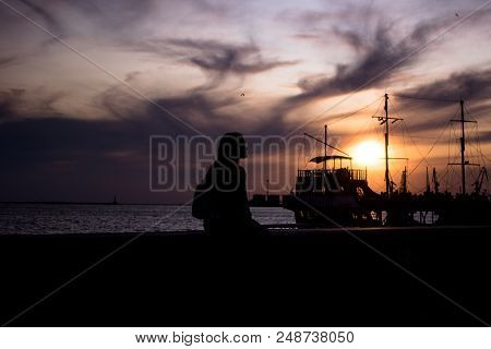 Silhouette Of A Girl On The Waterfront. Silhouette Of An Old Ship In The Background. The Girl Is Wai
