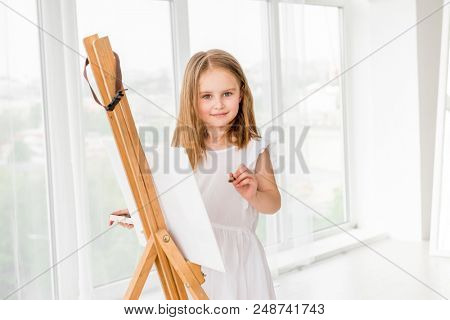 Pretty girl with brush in hand. Creative teen girl paitning a picture on easel. Girl practising drawing in art school