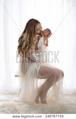 A Beautiful Mother Kissing Her Sleeping Newborn Daughter. She Is Wearing A Sheer Robe And Sitting In
