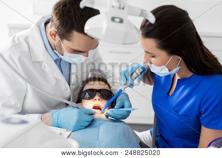medicine, dentistry and healthcare concept - dentist and assistant with dental drill and saliva ejector treating kid patient teeth at dental clinic