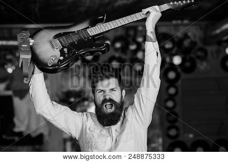 Man With Beard And Mustache On Shouting Face Holds Guitar Above His Head, Music Club Background. Roc