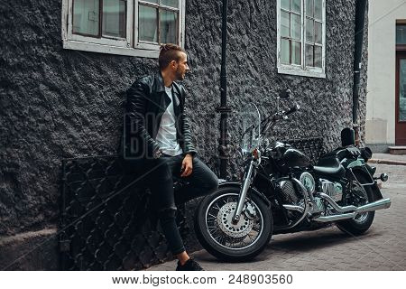 Fashionable Biker Dressed In A Black Leather Jacket And Jeans Leaning On A Wall Near His Retro Motor