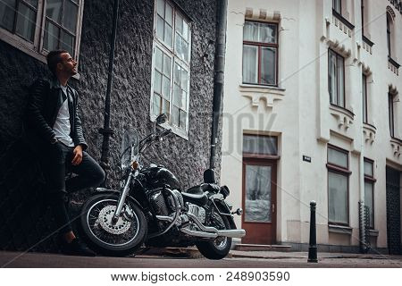 Fashionable Biker Dressed In A Black Leather Jacket And Jeans Leaning On A Wall Near His Retro Motor