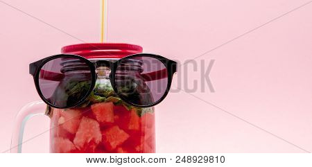 Glasses Lying On A Jar Of Cold Fresh Lemonade With Piece Of Watermelon And Ice On Pink Background.