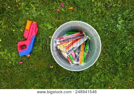 Summertime Fun Represented By A Bucket Full Of Popsicles And A Water Gun On The Grass