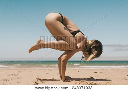 Side View Of Young Woman In Black Bikini Practicing Bakasana Yoga Position On Sandy Beach