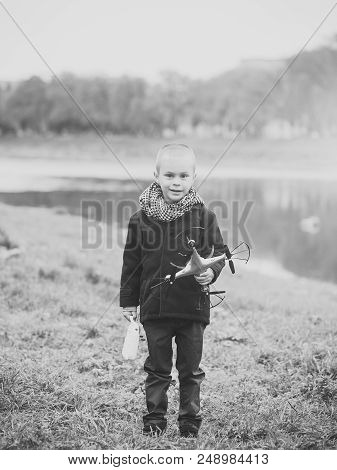 Child With Quadrupter In Autumn Scarf And Coat Standing At River Water On Green Grass On Natural Bac