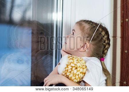 A Little Girl With A Toy Sits On The Windowsill And Draws On The Window.
