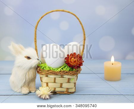 The Easter Bunny Sits Next To A Basket Full Of Eggs, Decorated With Dried Flowers. Burning Candles I
