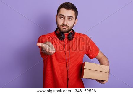 Horizontal Indoor Shot Of Handsome Hard Working Delivery Man Putting Hand Forward, Waiting For Money