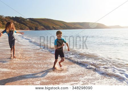 Two Children Wearing Wetsuits Running Through Waves On Summer Beach Vacation