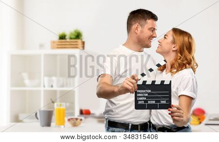 production, filmmaking and entertainment concept - portrait of happy couple in white t-shirts with clapperboard over home kitchen background