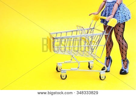 An Empty Supermarket Basket On A Yellow Background And A Toy Girl Shopper With Long Legs In High-hee