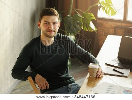 Smiling Freelancer Guy At Laptop Holding Coffee Cup Looking At Camera Sitting At Workdesk In Modern 