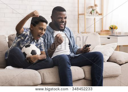 Afro Father And Son Soccer Fans Watching Football On Tv At Home And Emotionaly Cheering, Sitting Wit