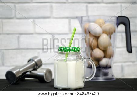 Protein Shake In A Mug Against The Background Of A Blender Bowl With Chicken Eggs And Dumbbells Agai