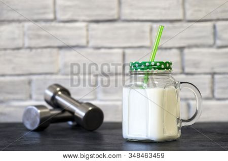 White Protein Cocktail In A Mug On The Background Of Dumbbells And A White Wall Decorated With Brick